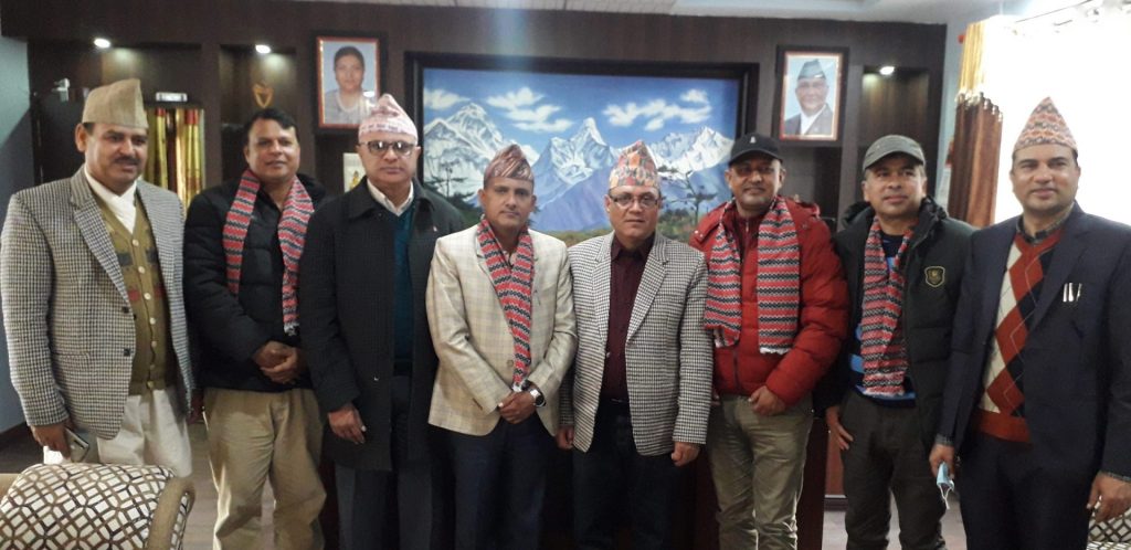 Group of eight men wearing traditional Nepali attire and scarves standing in a room with framed pictures and a mountain landscape painting, likely attending an event or meeting.