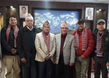 Group of eight men wearing traditional Nepali attire and scarves standing in a room with framed pictures and a mountain landscape painting, likely attending an event or meeting.
