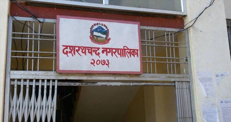 A close-up photograph of a signboard with a logo and text in Nepali script. The signboard reads 'Dasharathchand Municipality 2073' and features a logo with a mountain range and a river. The sign is white with a red border and is attached to a building with metal grills on the windows.