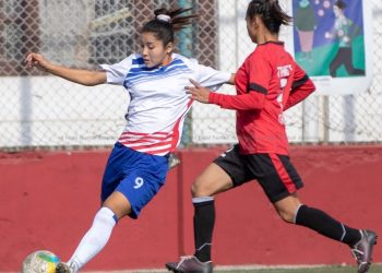 Two female soccer players in action on the field, one wearing a white and blue jersey with the number 9 and blue shorts, the other in a red jersey with black shorts, both with their hair tied back, playing with a multicolored soccer ball.