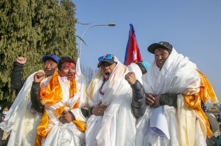 Group of men in traditional attire, wrapped in white and yellow cloths, with red tika on their foreheads, shouting and raising fists in celebration or protest, with a Nepali flag visible in the background.