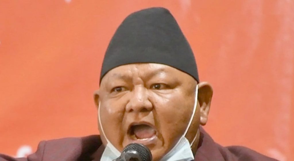 Close-up of a Nepali man wearing a black Dhaka topi and a maroon jacket, speaking into a microphone. He appears to be in the middle of a passionate speech, mouth open and eyes focused. The background is a vibrant orange color.