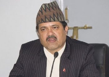 Portrait of Mayor Baldev Singh Gomden Tamang wearing a traditional Nepali Dhaka topi and a black pinstripe suit, seated in a black leather chair with a microphone in front of him, in a formal setting with a plain white wall and a brass tap in the background.