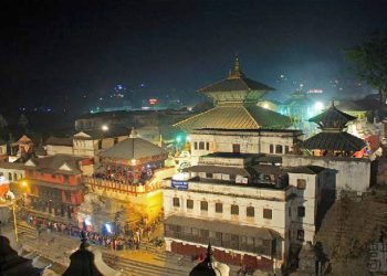 A nighttime view of a densely packed cluster of traditional Nepalese buildings with pagoda-style roofs, illuminated by vibrant lights. The main structure in the center has a prominent golden roof and white walls. People are visible on the steps and balconies. The scene is set in a bustling urban area with a stone staircase leading up to the buildings.