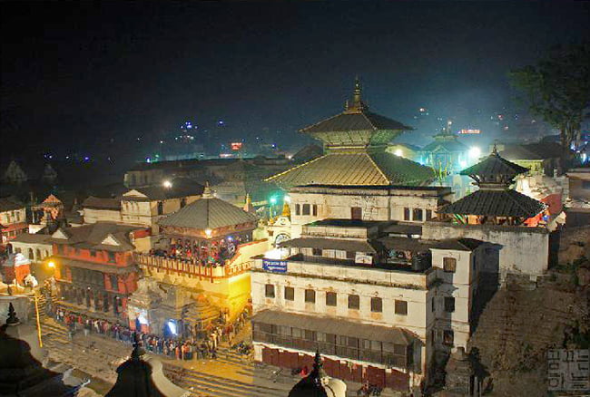 A nighttime view of a densely packed cluster of traditional Nepalese buildings with pagoda-style roofs, illuminated by vibrant lights. The main structure in the center has a prominent golden roof and white walls. People are visible on the steps and balconies. The scene is set in a bustling urban area with a stone staircase leading up to the buildings.