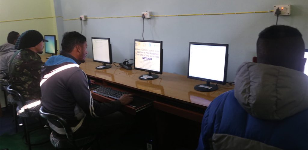 Four men are sitting at a row of computer desks, each using a computer. The man on the left is wearing a black beanie and a camouflage jacket. The man next to him is wearing a gray and white jacket. The man on the right is wearing a blue jacket with a gray hood. The fourth man is partially visible on the far left. They are in a room with light blue walls and a wooden desk.