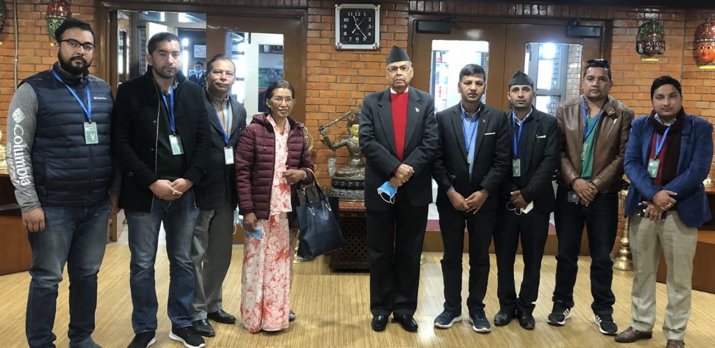 Group of eight individuals standing in a line, posing for a photograph. They are wearing formal and semi-formal attire, with some sporting blue lanyards. The setting appears to be an indoor space with brick walls, wooden floors, and decorative elements.