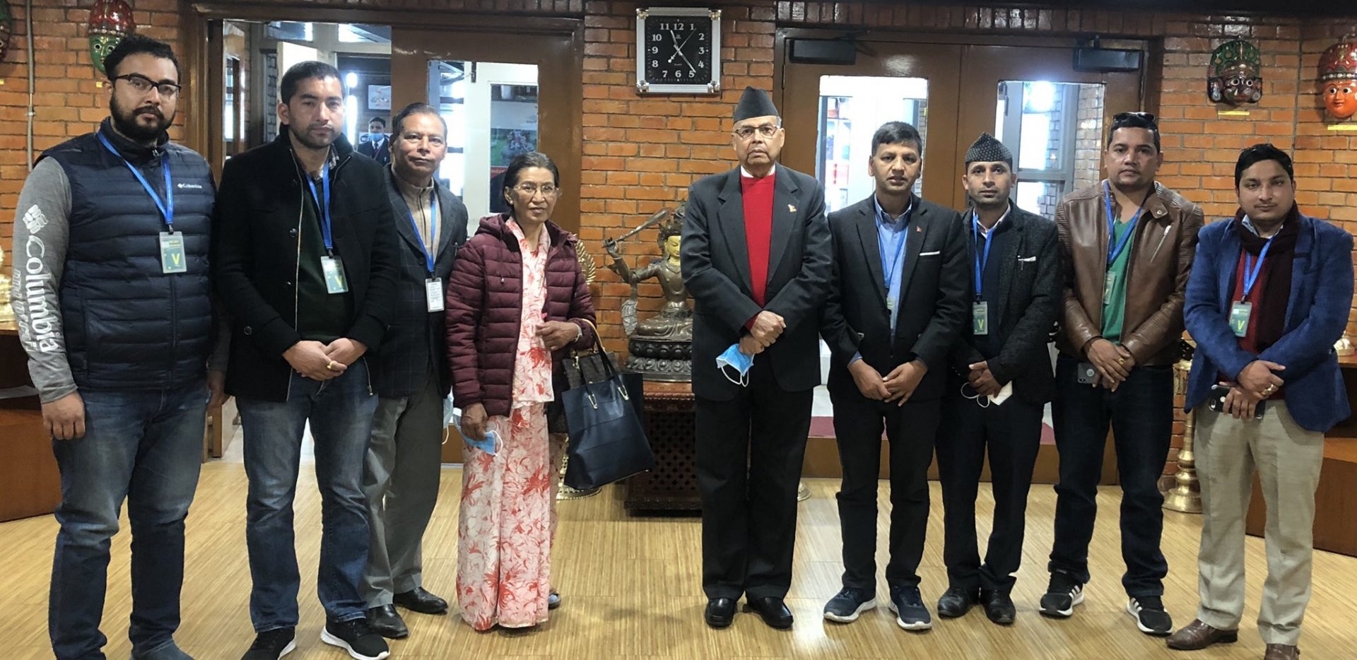Group of eight individuals standing in a line, posing for a photograph. They are wearing formal and semi-formal attire, with some sporting blue lanyards. The setting appears to be an indoor space with brick walls, wooden floors, and decorative elements.