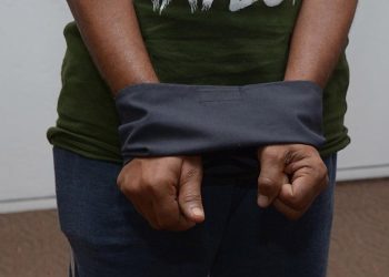 A person wearing a green t-shirt and blue jeans with their hands cuffed behind their back with a dark cloth, standing in front of a white wall and brown carpeted floor.