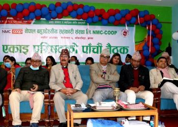 Group of men and women sitting on couches in front of a banner at a meeting. The men are wearing traditional Nepali attire including Dhaka topi hats. Specific individuals visible are (from left to right): an older man in a dark green jacket and grey pants, another man in a dark jacket and white pants, a man in a tan suit with a red shirt, a man in a tan jacket with his arms crossed, a man in a black jacket and white pants, a man in a tan jacket and light grey pants, and a man in a dark jacket and blue jeans. They appear to be at a formal event or meeting, possibly at a cooperative or community center. The setting is indoors with green walls, blue and red balloons, and a large white banner in Nepali script.