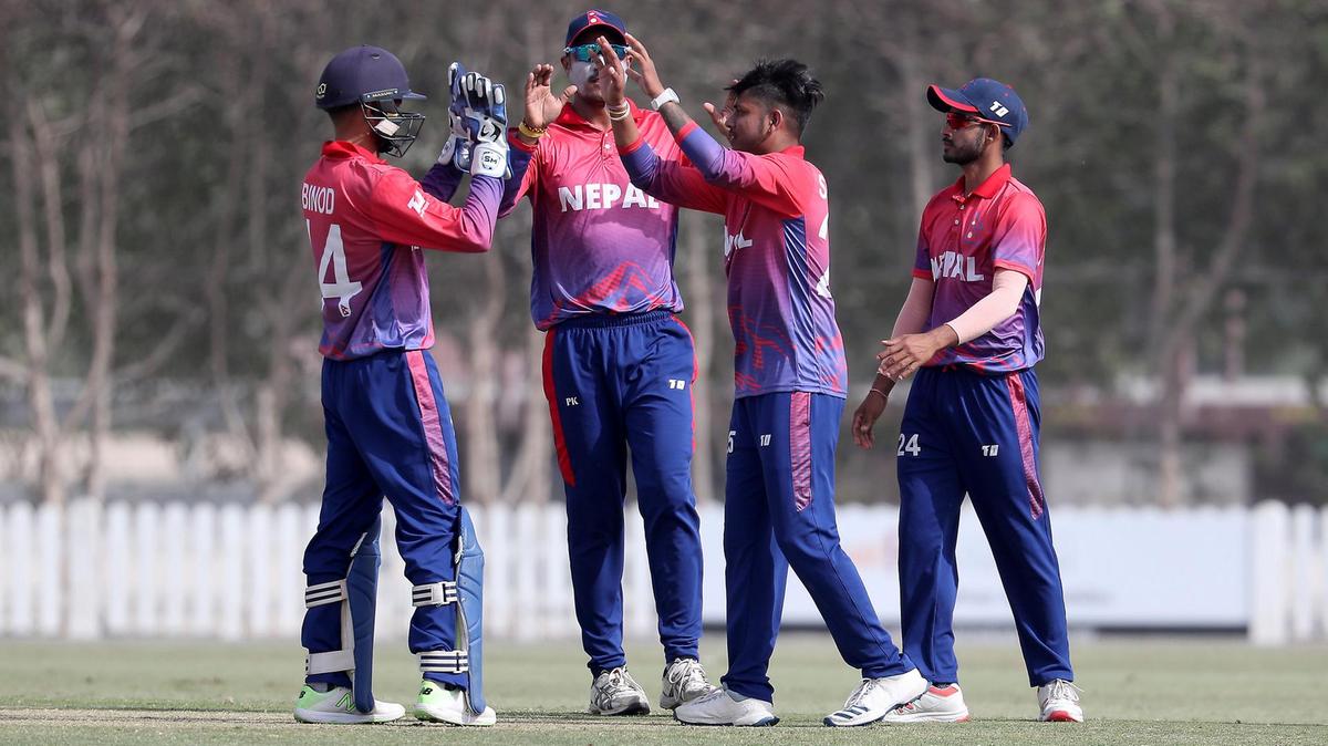 Four Nepali cricket players in red and blue uniforms with 'NEPAL' on their jerseys are celebrating on the field. The player on the left is wearing a helmet and gloves, while the others have their hands raised in a high-five. The players are standing on a green field with trees and a white fence in the background.