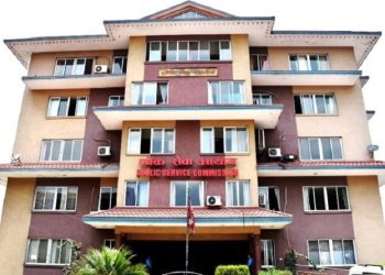 The Public Service Commission building with a red sign in English and Nepali, multiple windows and balconies, air conditioners, and a flagpole with a red flag, located in a urban setting with trees and bushes in front.
