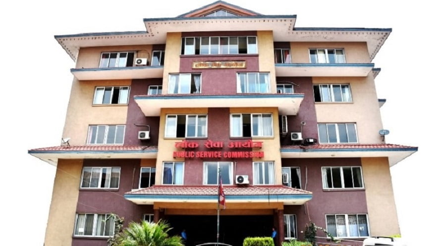 The Public Service Commission building with a red sign in English and Nepali, multiple windows and balconies, air conditioners, and a flagpole with a red flag, located in a urban setting with trees and bushes in front.