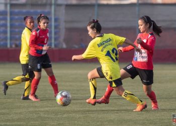 Four women in soccer uniforms playing on a green field. A woman in a yellow jersey with 'BIRATHNAGA' and '13' on the back is kicking the ball while being challenged by a woman in a red jersey. Two other women in red and yellow jerseys are nearby.