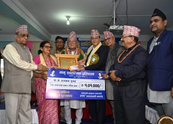 Prof. Dr. Jagaman Gurung is being honored with a framed certificate and a large cheque for NPR 109,000 by multiple dignitaries, including a woman in a pink dress, inside a decorated indoor setting, with all individuals wearing traditional Nepali attire and accessories.