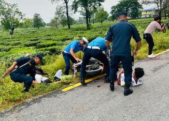 Security personnel in uniform attending to people lying on the roadside near a tea garden, with a motorbike nearby, in what appears to be an accident scene.