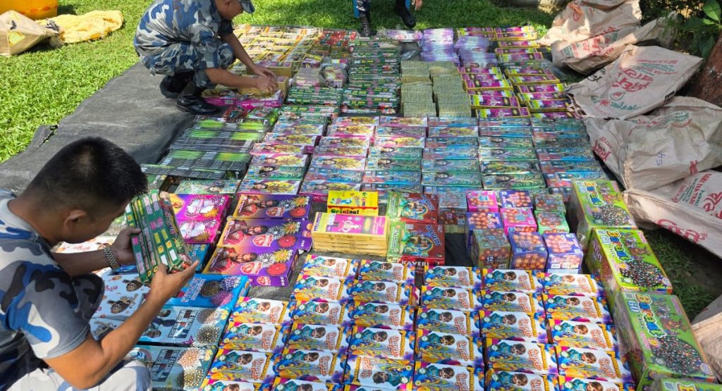 Detailed image of seized fireworks display, with two Armed Police Force personnel in camouflage uniforms arranging and inspecting colorful boxes of explosives, including various types of crackers and pyrotechnics, on a black tarp in a grassy area, with jute bags on the right, under natural daylight.