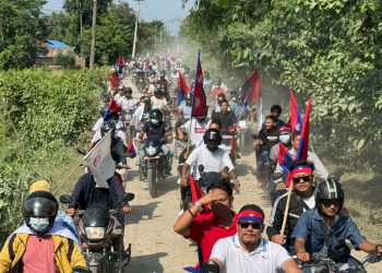 A large group of motorcyclists, including Khagendra Raj Angdembe, Asha Gurung, and Haricharan Shivakoti, participate in a rally on a dirt road in Virtamod, Jhapa. They are holding red and blue flags and wearing face masks. The scene is set against a backdrop of lush greenery and trees, with a few buildings visible in the distance. The atmosphere appears to be one of protest or demonstration.