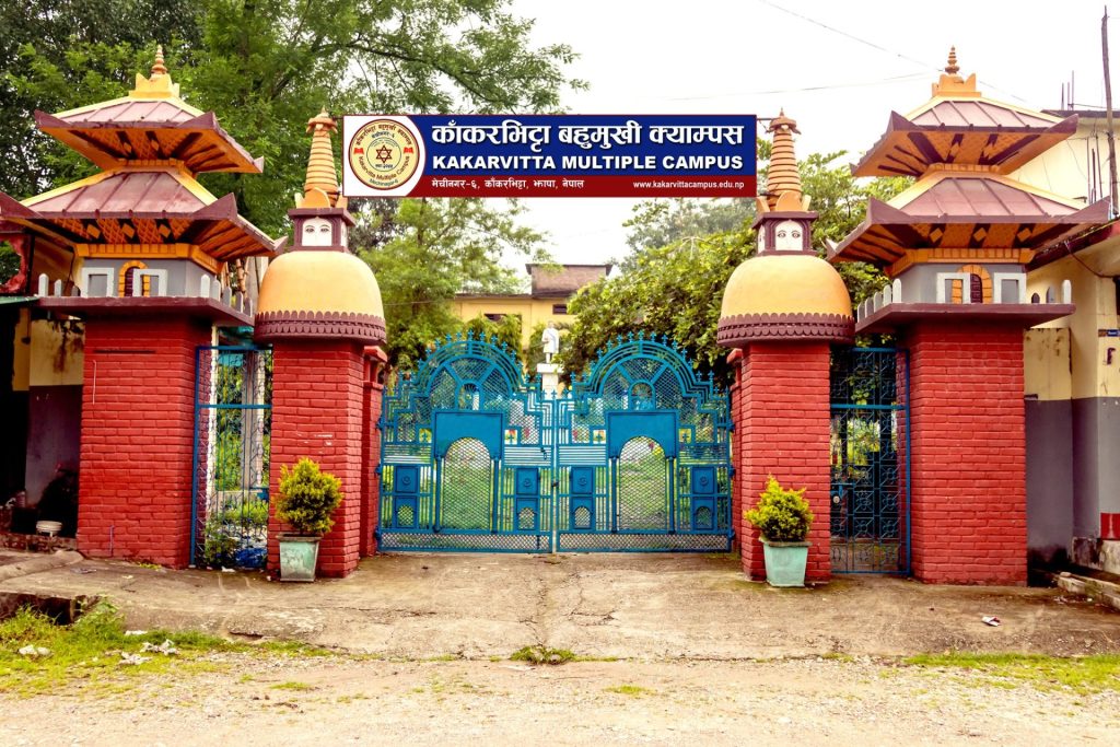 Kakarbhitta Multiple Campus entrance gate with blue metal doors and red brick pillars, surrounded by green trees and a cloudy sky.