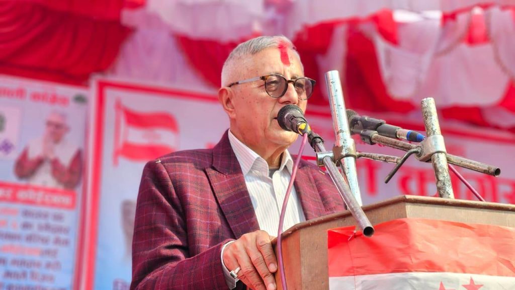 Dr. Shekhar Koirala, a middle-aged man with short gray hair and glasses, wearing a red plaid jacket, speaking into a microphone at a podium with a red and white cloth draped over it, in front of a red and white decorated background.