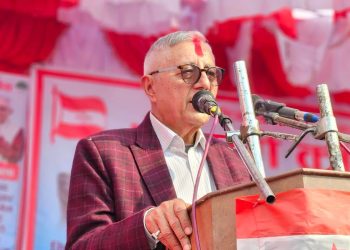 Dr. Shekhar Koirala, a middle-aged man with short gray hair and glasses, wearing a red plaid jacket, speaking into a microphone at a podium with a red and white cloth draped over it, in front of a red and white decorated background.
