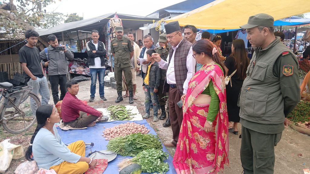 Mayor Gopal Chandra Budhathoki and Deputy Mayor Mina Pokhrel Upreti inspecting the Dhulabari market with officials and local vendors. They are standing near a table with vegetables and potatoes. The scene includes several people in casual and formal attire, with some taking pictures or observing.