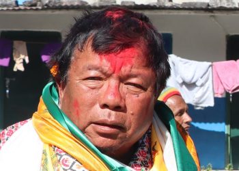 Close-up of Dr. Mahavir Pun with red tika on his forehead, wearing a white, green, and yellow shawl, standing in front of a house with clothes hanging outside, looking downwards with a calm expression.