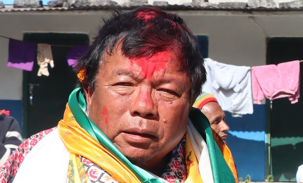 Close-up of Dr. Mahavir Pun with red tika on his forehead, wearing a white, green, and yellow shawl, standing in front of a house with clothes hanging outside, looking downwards with a calm expression.