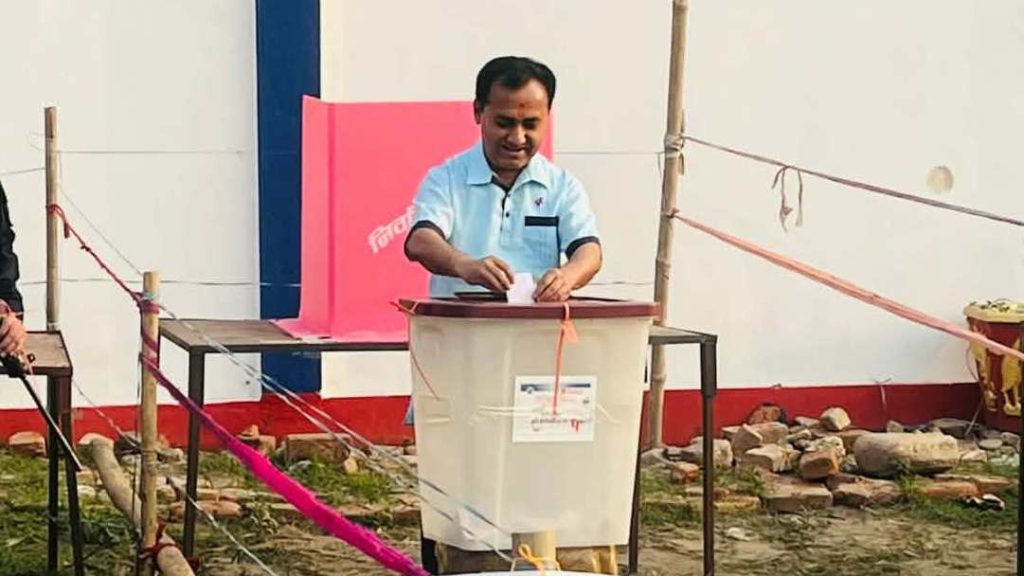 A man in a light blue polo shirt is inserting a ballot paper into a large white box with a brown lid, likely a polling station during an election. The box has a label on it and is secured with orange straps. The man has short black hair and appears to be focused on the task. He is standing in a semi-outdoor setting with a white wall behind him, marked with red and blue stripes at the bottom. A pink barrier tape is visible in the foreground.
