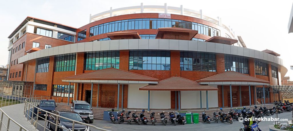 The image shows a large, modern building with a curved facade and numerous windows. The building appears to be a government office or institutional facility, possibly related to passport services. Several motorcycles are parked in front of the building, along with a few cars. The scene is set during the daytime with a clear sky in the background.