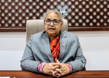 Prime Minister Sushila Karki sitting at a desk with her hands clasped together, wearing a gray blazer and an orange scarf, seated in a beige chair with a wooden frame and lattice design on the wall behind her.