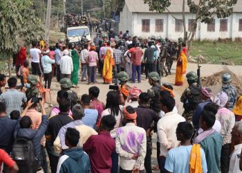 A large crowd of people, including Nepali Congress and Rastriya Swatantra Party supporters, are gathered outside a polling station in Balara, Sarlahi, amidst a dispute between the two parties' workers, with some individuals wearing green military helmets and others holding phones to capture the scene, as security personnel try to maintain order.