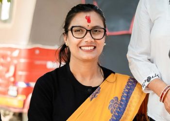 Sobita Gautam, a prominent leader of Rastriya Swatantra Party (Raswapa), smiling and wearing a black top with a yellow sari featuring blue designs, adorned with a red tika on her forehead, standing in front of a red and white background.