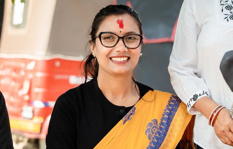 Sobita Gautam, a prominent leader of Rastriya Swatantra Party (Raswapa), smiling and wearing a black top with a yellow sari featuring blue designs, adorned with a red tika on her forehead, standing in front of a red and white background.