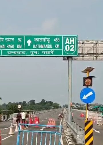 A roadblock at the Nepal-India border with a green signpost indicating directions to Dhulabari, Singalila National Park, and Kathmandu. The road is closed with barriers, cones, and a 'STOP' sign. No specific people are visible.