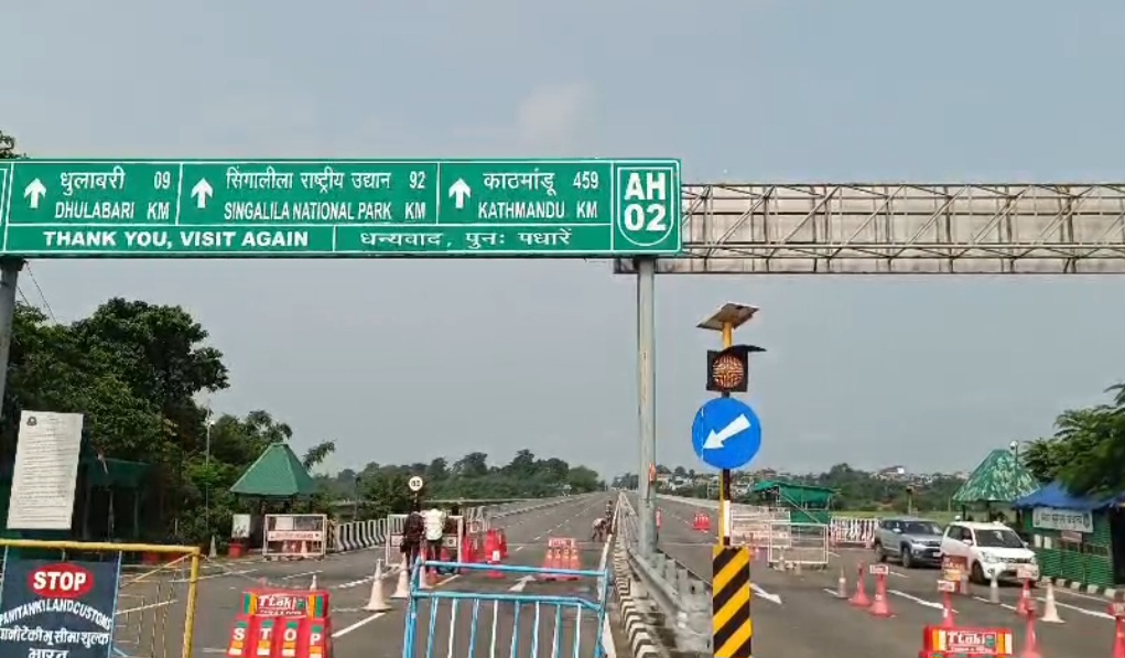 A roadblock at the Nepal-India border with a green signpost indicating directions to Dhulabari, Singalila National Park, and Kathmandu. The road is closed with barriers, cones, and a 'STOP' sign. No specific people are visible.