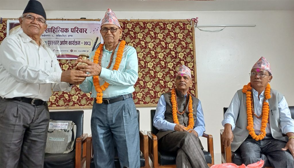 HONORING TRADITION: Renowned Nepali literary figures, from left, HOM SUWEDI, CHUDA MANI WASHISHT, GANGA PRASAD OFFICIAL, and another distinguished guest adorned in traditional Nepali attire, including Dhaka topi hats and marigold garlands, participate in a ceremonial event. They are seated and standing in a formal setting with a decorative backdrop, likely during a cultural or literary ceremony in Jhapa, Nepal.