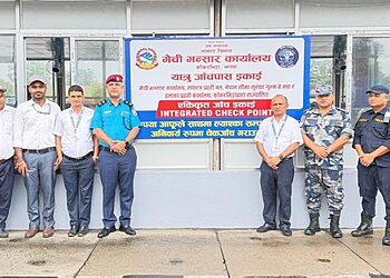 Group photo of officials from various agencies standing in front of a signboard for the Integrated Check Point at Kakarbhitta border crossing. Visible are (from left to right): a security officer in camouflage uniform, officials in plain clothes, and uniformed personnel from Nepal Police, Armed Police Force, and Nepal Customs. They are standing in front of a large sign with Nepali and English text. The scene is set on a concrete pavement outside a building with glass windows.
