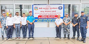 Group photo of officials from various agencies standing in front of a signboard for the Integrated Check Point at Kakarbhitta border crossing. Visible are (from left to right): a security officer in camouflage uniform, officials in plain clothes, and uniformed personnel from Nepal Police, Armed Police Force, and Nepal Customs. They are standing in front of a large sign with Nepali and English text. The scene is set on a concrete pavement outside a building with glass windows.