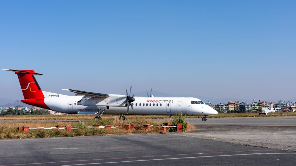 A white and red Shree Airlines plane, registration number 9N-ANE, taxis on the runway with a cityscape and mountains in the background.