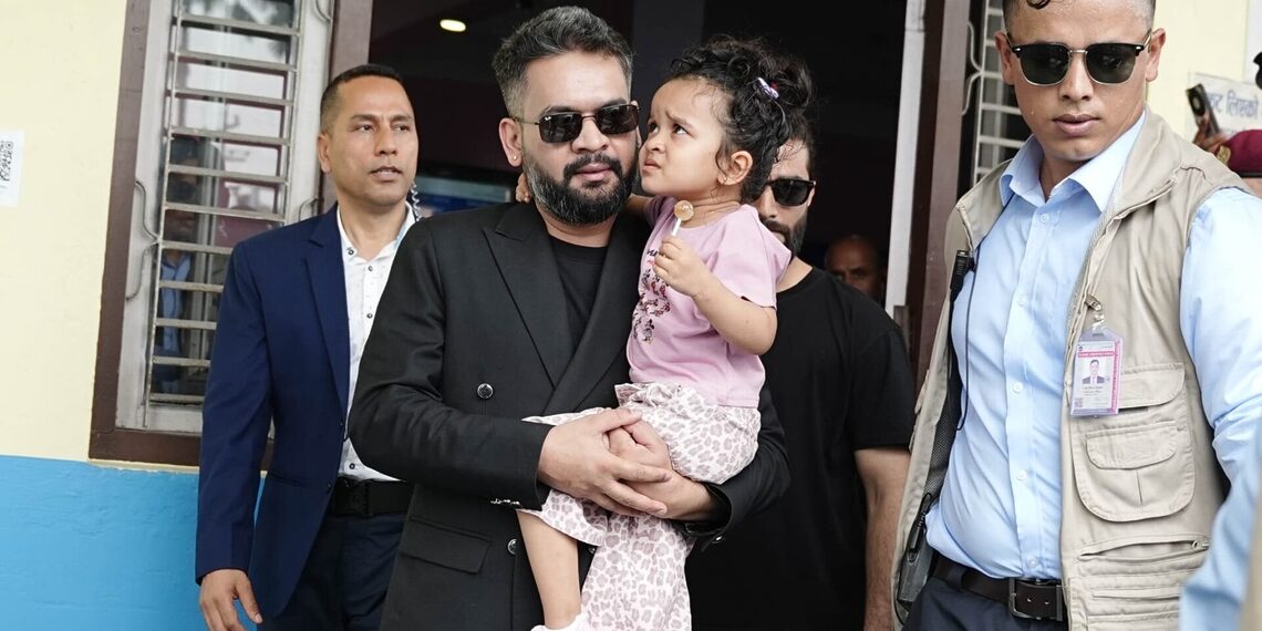 Prime Minister Balen Shah, wearing a black suit and sunglasses, holds a young girl in a pink dress. He is flanked by two men in sunglasses, one in a blue suit and the other in a tan vest. They stand in front of a building with open windows.