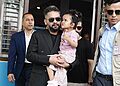 Prime Minister Balen Shah, wearing a black suit and sunglasses, holds a young girl in a pink dress. He is flanked by two men in sunglasses, one in a blue suit and the other in a tan vest. They stand in front of a building with open windows.
