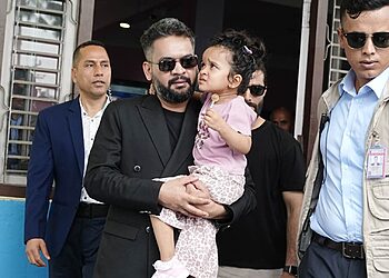Prime Minister Balen Shah, wearing a black suit and sunglasses, holds a young girl in a pink dress. He is flanked by two men in sunglasses, one in a blue suit and the other in a tan vest. They stand in front of a building with open windows.
