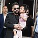 Prime Minister Balen Shah, wearing a black suit and sunglasses, holds a young girl in a pink dress. He is flanked by two men in sunglasses, one in a blue suit and the other in a tan vest. They stand in front of a building with open windows.