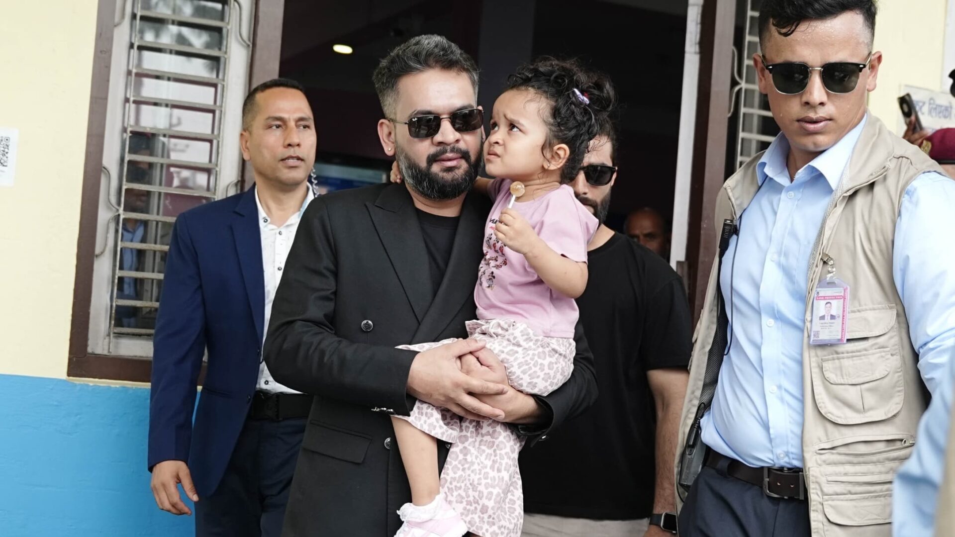 Prime Minister Balen Shah, wearing a black suit and sunglasses, holds a young girl in a pink dress. He is flanked by two men in sunglasses, one in a blue suit and the other in a tan vest. They stand in front of a building with open windows.