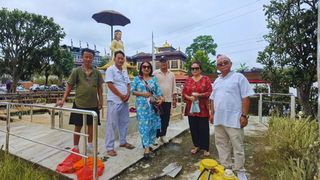 A group of seven people, including Harkabahadur Fiyack Limbu, are standing near a newly installed full-size statue of King Baliraj in Shrijangapark, Kakarbhitta, Mechinagar. The statue depicts King Baliraj with a bowl of coins in his hand. The group is posing in front of a Buddhist-style building and a large golden statue. The scene is set on a cloudy day with trees and a metal bridge in the background.