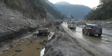 Vehicles navigate through a damaged section of BP Highway with excavators working on the side, muddy water flowing through the roadside, and mountains in the background. No specific people are visible.