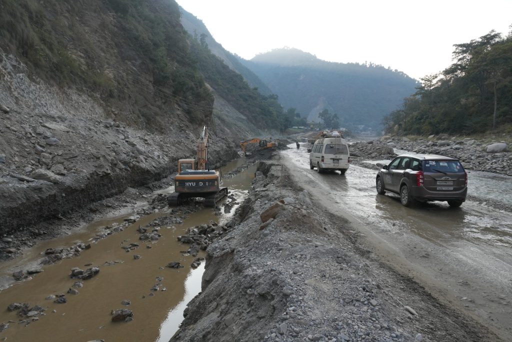 Vehicles navigate through a damaged section of BP Highway with excavators working on the side, muddy water flowing through the roadside, and mountains in the background. No specific people are visible.