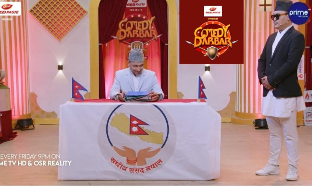 The image shows a scene from the Nepali comedy show 'Comedy Darbar'. A man dressed in a white suit and grey hat sits at a table with the logo of the Nepali parliament on it, holding some papers. Another man in a black suit, white pants, and a hat stands to his right. The setting appears to be a TV studio with a red and gold backdrop.