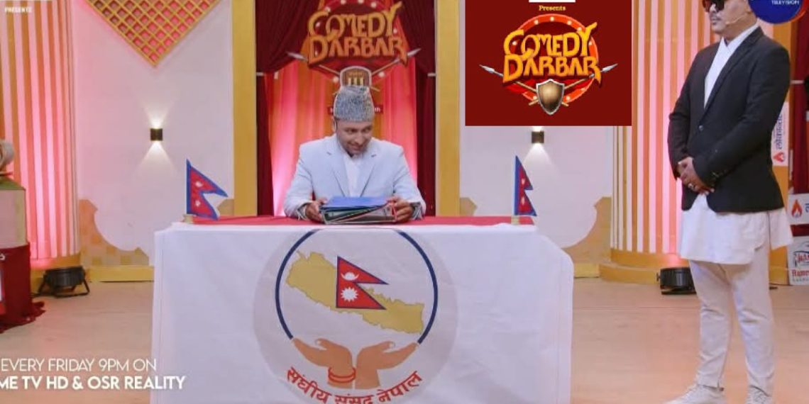 The image shows a scene from the Nepali comedy show 'Comedy Darbar'. A man dressed in a white suit and grey hat sits at a table with the logo of the Nepali parliament on it, holding some papers. Another man in a black suit, white pants, and a hat stands to his right. The setting appears to be a TV studio with a red and gold backdrop.